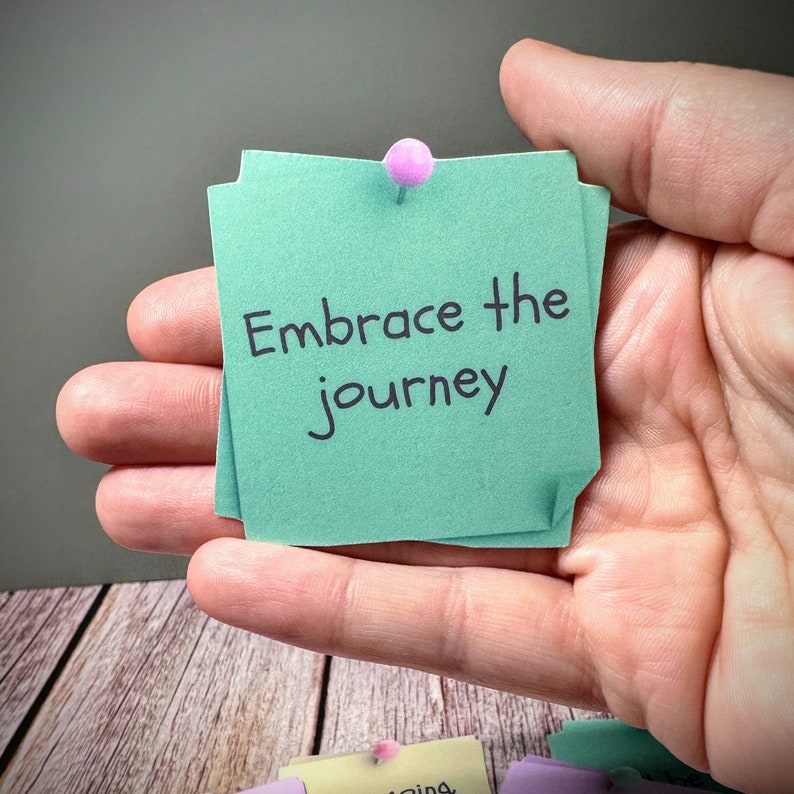 Puede incluir: Una nota adhesiva verde con el texto "Embrace the journey" escrito en marcador negro. La nota est&aacute; fijada a una mano con un chinche rosa.