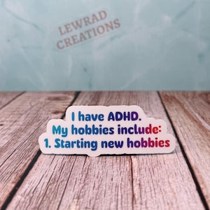 May include: A white sticker with the text "I have ADHD. My hobbies include: 1. Starting new hobbies" in blue and red lettering. The sticker has a rounded shape and is on a wooden surface.