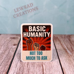 May include: A square sticker with the words "BASIC HUMANITY" in bold white letters. Below, it reads "RESPECT, DIGNITY, COMPASSION". The bottom text says "NOT TOO MUCH TO ASK". The design features three raised fists.