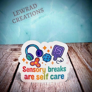 May include: A sticker with the text "Sensory breaks are self care" in rainbow colors. The sticker features illustrations of headphones, a fidget spinner, a textured ball, and a game controller. The sticker has a white border and the brand name "LEWRAD CREATIONS" is visible.