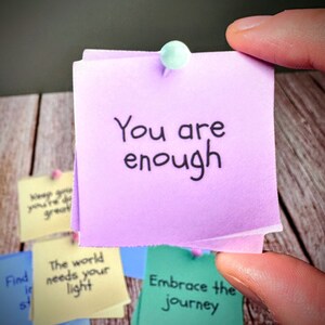 Puede incluir: Una nota adhesiva morada con el texto "You are enough" escrito en marcador negro. La nota est&aacute; fijada a una superficie de madera con otras notas adhesivas de colores.