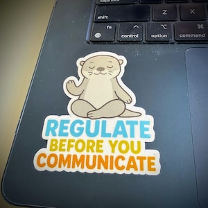Puede incluir: Una pegatina troquelada que muestra una nutria de dibujos animados de color marr&oacute;n claro sentada en posici&oacute;n de meditaci&oacute;n con las piernas cruzadas y los ojos cerrados. Debajo, el texto dice REGULATE en azul, BEFORE YOU en amarillo y COMMUNICATE en naranja.