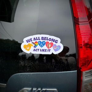 May include: A sticker with the text "WE ALL BELONG ACT LIKE IT" and colorful heart designs representing inclusivity. The sticker is affixed to a vehicle's rear window, promoting diversity and acceptance.