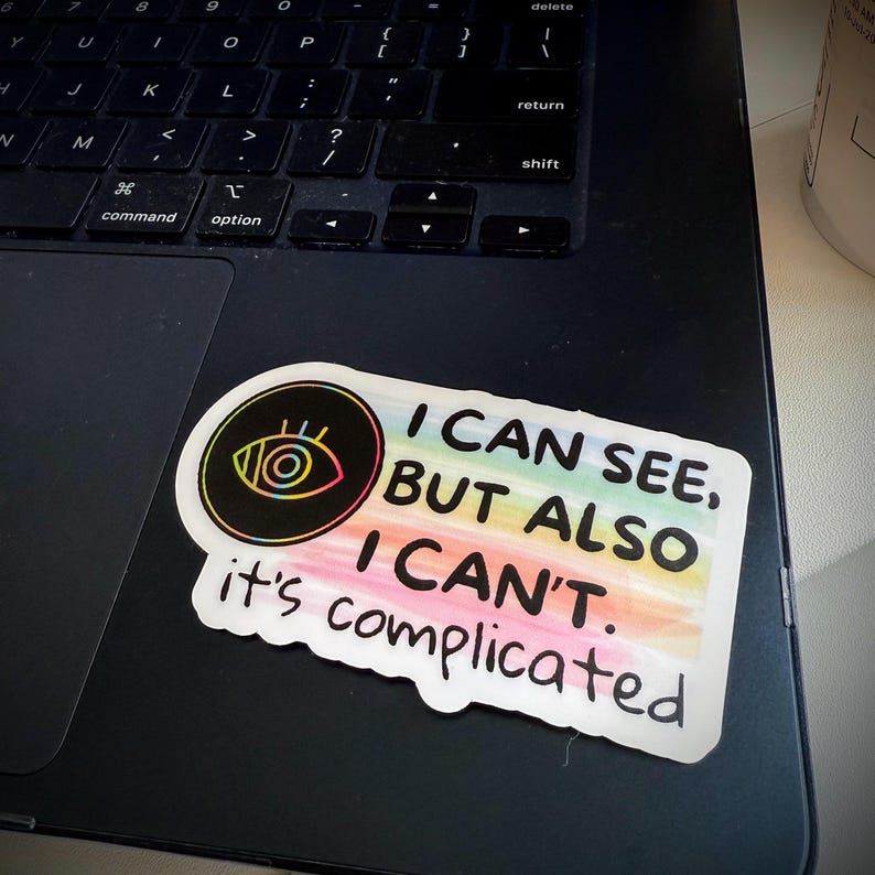 May include: A sticker with the text "I CAN SEE, BUT ALSO I CAN'T. it's complicated" in black font. The sticker has a rainbow gradient background and a circular graphic of an eye with a black outline.