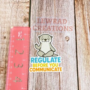 Puede incluir: Una pegatina troquelada que muestra una nutria de dibujos animados de color marr&oacute;n claro sentada en posici&oacute;n de loto. Debajo de la nutria, el texto dice REGULATE en azul y BEFORE YOU COMMUNICATE en amarillo. Una regla rosa mide unos 10 cm.
