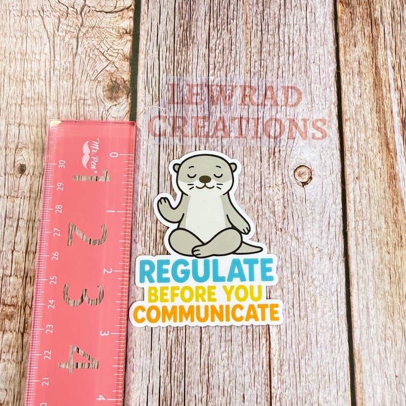 Puede incluir: Una pegatina troquelada que muestra una nutria de dibujos animados de color marr&oacute;n claro sentada en posici&oacute;n de loto. Debajo de la nutria, el texto dice REGULATE en azul y BEFORE YOU COMMUNICATE en amarillo. Una regla rosa mide unos 10 cm.