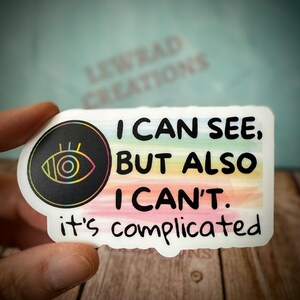May include: A white sticker with the text "I CAN SEE, BUT ALSO I CAN'T. it's complicated." The sticker has a black circle with an eye design and a rainbow gradient background.