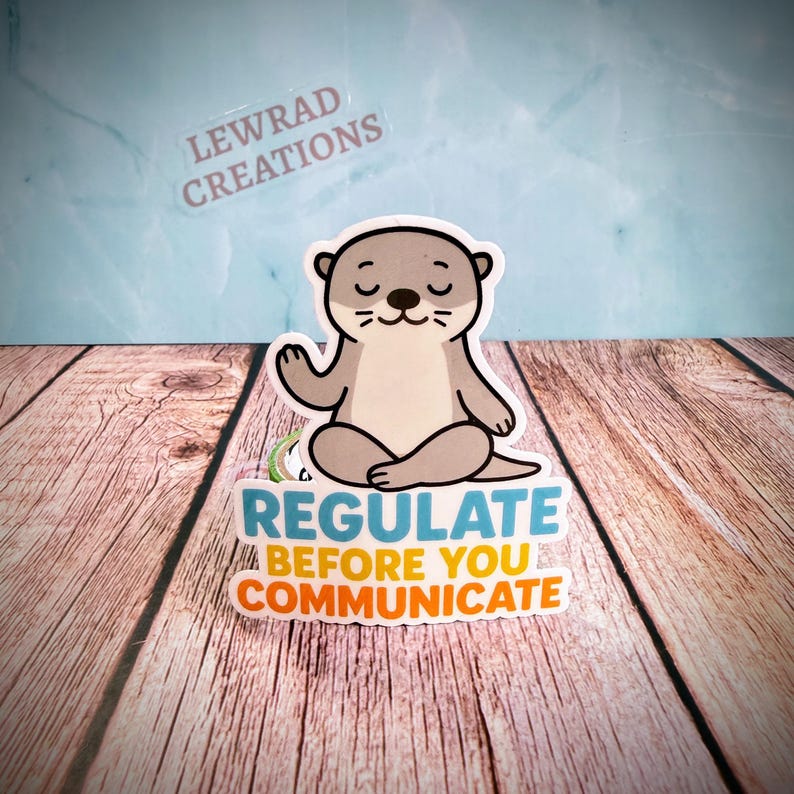 Puede incluir: Una pegatina troquelada que muestra una nutria de dibujos animados de color marr&oacute;n claro sentada en posici&oacute;n de meditaci&oacute;n con las piernas cruzadas y los ojos cerrados. Debajo de la nutria, el texto dice REGULATE en azul, BEFORE YOU en amarillo y COMMUNICATE en naranja. El nombre de la marca LEWRAD CREATIONS es visible al fondo.