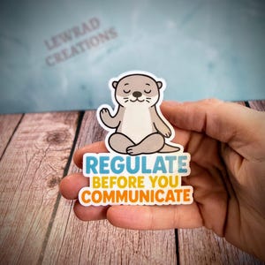 Puede incluir: Una pegatina troquelada que muestra una nutria de dibujos animados de color marr&oacute;n claro sentada en posici&oacute;n de meditaci&oacute;n con los ojos cerrados. Debajo, el texto dice REGULATE en azul, BEFORE YOU en amarillo y COMMUNICATE en naranja, en negrita.