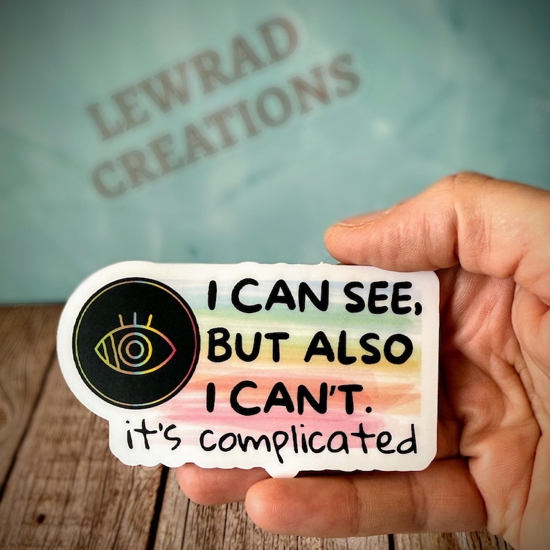 May include: A white sticker with the text "I CAN SEE, BUT ALSO I CAN'T. it's complicated." The sticker has a black circle with an eye design and a rainbow-colored background.