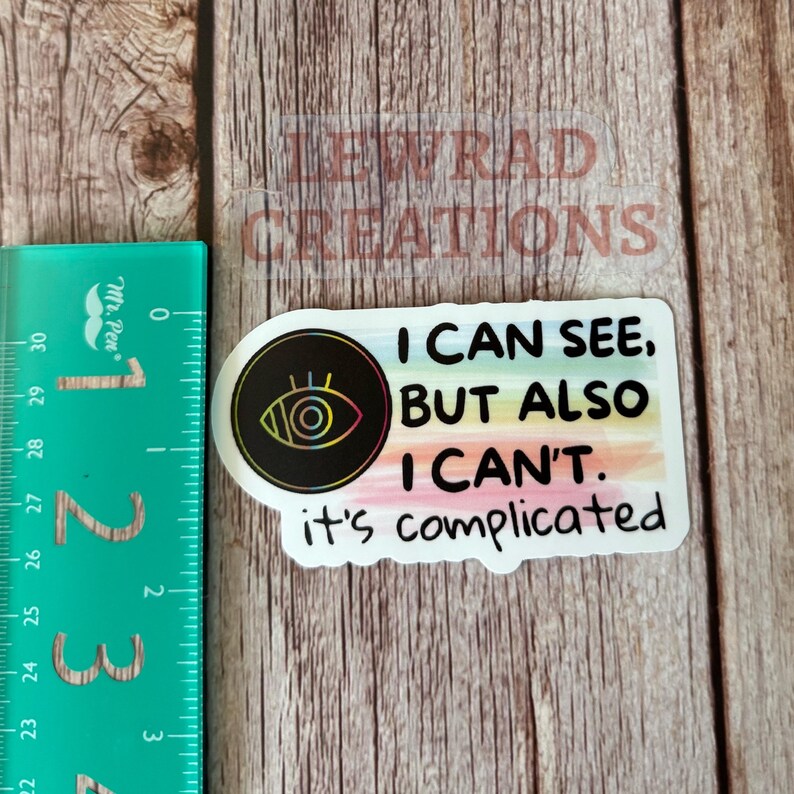 May include: A sticker with the text "I CAN SEE, BUT ALSO I CAN'T. it's complicated" in black font. The sticker has a black circle with an eye design. A green ruler is on the left.