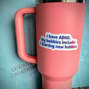 May include: A large, coral-colored tumbler with a handle and a silver rim. A sticker on the side reads "I have ADHD. My hobbies include: 1. Starting new hobbies." The tumbler has a clear lid and is made by Lewral Creations.