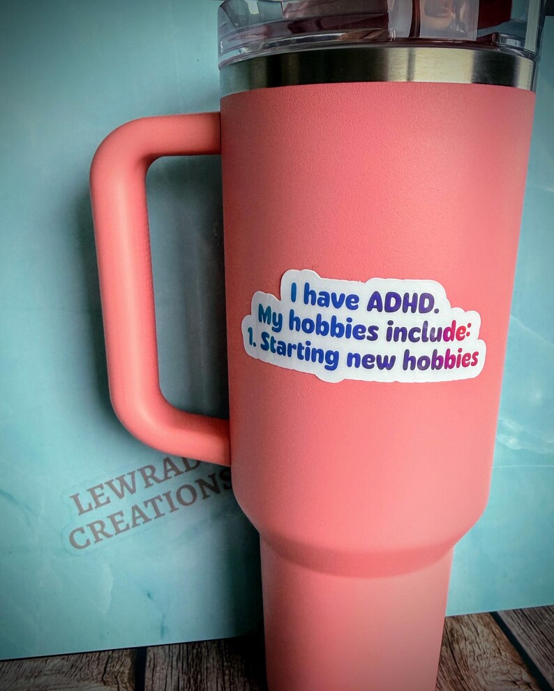 May include: A large, coral-colored tumbler with a handle and a silver rim. A sticker on the side reads "I have ADHD. My hobbies include: 1. Starting new hobbies." The tumbler has a clear lid and is made by Lewral Creations.