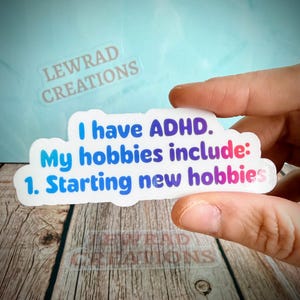 May include: A white sticker with the text "I have ADHD. My hobbies include: 1. Starting new hobbies" in blue and pink lettering. The sticker is held by a person's fingers. The sticker has the words "LEWRAD CREATIONS" printed on it.