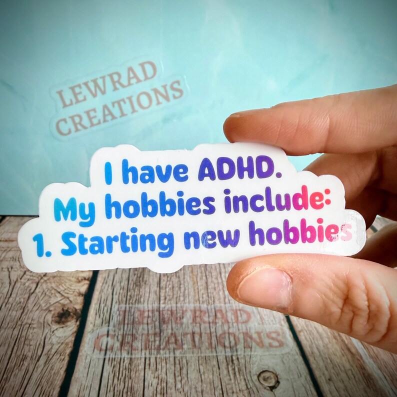 May include: A white sticker with the text "I have ADHD. My hobbies include: 1. Starting new hobbies" in blue and pink lettering. The sticker is held by a person's fingers. The sticker has the words "LEWRAD CREATIONS" printed on it.