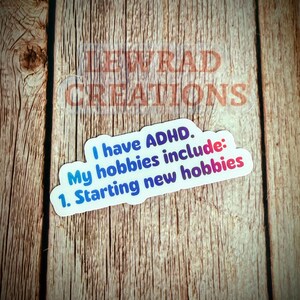 May include: A white sticker with a blue and purple gradient border, featuring the text "I have ADHD. My hobbies include: 1. Starting new hobbies." The sticker is on a wooden surface, with the text "LEWRAD CREATIONS" faintly visible.