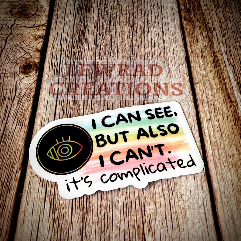 May include: A sticker with the text "I CAN SEE, BUT ALSO I CAN'T. it's complicated." The sticker features a black circle with an eye design and a rainbow-colored background. The sticker is on a wooden surface.