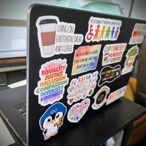 Pu&ograve; includere: Un laptop nero ricoperto di adesivi colorati. Gli adesivi includono frasi come "Living on Existential Dread and Coffee", "Equality, Justice, Inclusion, Compassion, Diversity, Kindness" e "Optimism Powered by Coffee". Altri adesivi presentano grafiche e testi relativi all'accessibilit&agrave; e all'autismo.