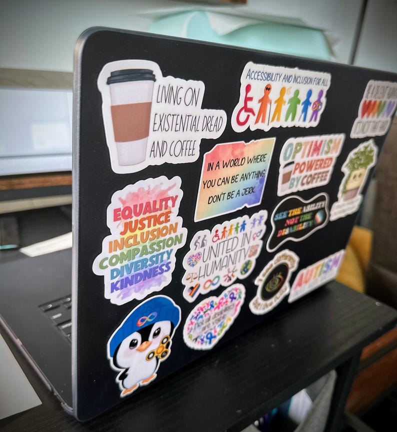 Pu&ograve; includere: Un laptop nero ricoperto di adesivi colorati. Gli adesivi includono frasi come "Living on Existential Dread and Coffee", "Equality, Justice, Inclusion, Compassion, Diversity, Kindness" e "Optimism Powered by Coffee". Altri adesivi presentano grafiche e testi relativi all'accessibilit&agrave; e all'autismo.