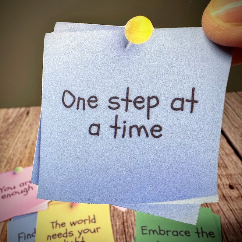 Puede incluir: Una nota adhesiva azul con el texto "One step at a time" escrito en marcador negro. La nota est&aacute; fijada a una superficie de madera con un chinche amarillo.