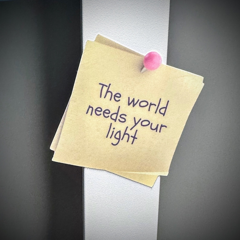 Puede incluir: Una nota adhesiva amarilla con el texto "The world needs your light" escrito en tinta negra. La nota est&aacute; pegada a una superficie blanca con un chinche rosa.