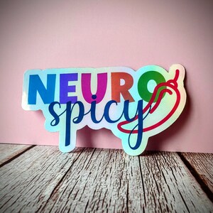 Pu&ograve; includere: Un adesivo olografico con le parole "NEURO spicy" in un font colorato. Un peperoncino rosso con uno stelo verde &egrave; accanto alla parola "spicy". L'adesivo &egrave; su una superficie di legno con uno sfondo rosa.