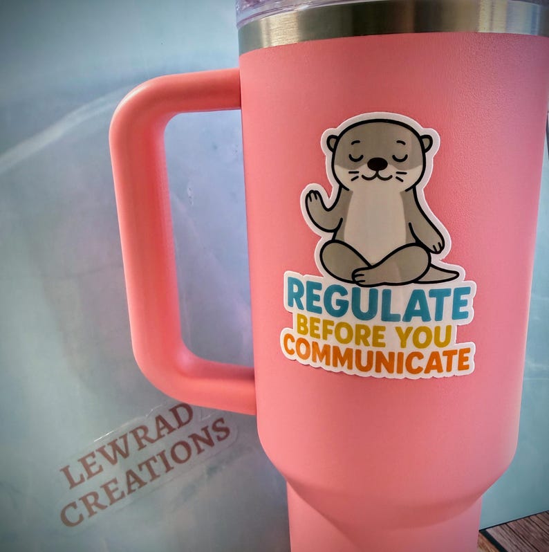 Puede incluir: Un vaso t&eacute;rmico de color rosa mate con asa y borde plateado. Una pegatina en la parte frontal muestra una nutria de dibujos animados sentada en posici&oacute;n de meditaci&oacute;n con el texto "REGULATE BEFORE YOU COMMUNICATE" en letras azules, amarillas y naranjas.