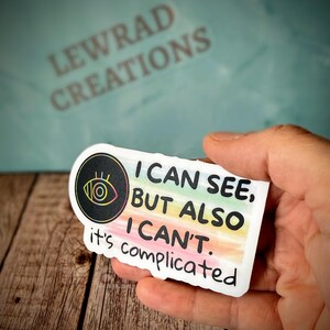 May include: A white sticker with the text "I CAN SEE, BUT ALSO I CAN'T. it's complicated." The sticker has a black circle with an eye graphic and rainbow stripes. The sticker is held in a hand.