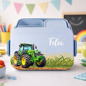 Könnte beinhalten: Hellblaue Brotdose mit einem grünen und gelben Traktor-Motiv und dem Namen "Felix" in weißer Schrift. Die Brotdose hat einen Griff und zwei Fächer. Der Traktor steht auf einem braunen Feld mit gelbem Gras.