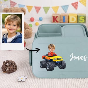 Könnte beinhalten: Hellblaue Brotdose mit einem Cartoon-Bild eines Jungen in einem Monstertruck und dem Namen "Jonas" in weißer Schrift. Die Brotdose hat drei Fächer und einen Deckel. Ein Foto eines Jungen befindet sich daneben.