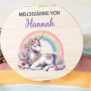 May include: A wooden tooth fairy box with a hinged lid, featuring a unicorn design with a rainbow. The box is open, revealing a circular interior with labeled tooth compartments. The text "MILCHZ&Auml;HNE VON Hannah" is on the lid.