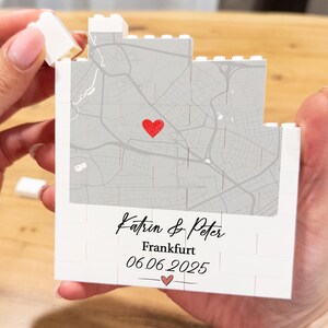 Pode incluir: Uma lembrança branca em forma de bloco com um design de mapa cinza e um coração vermelho marcando um local. O texto "Katrin & Peter", "Frankfurt" e a data "06.06.2025" estão impressos no bloco. A lembrança é segurada por uma pessoa.