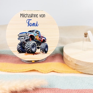 Könnte beinhalten: Eine Holz-Andenkenbox mit Scharnierdeckel und einer kleinen Glasflasche. Eine runde Holzplakette mit dem Text "Milchzähne von Toni" und einer farbenfrohen Monster-Truck-Illustration ist ebenfalls vorhanden. Der Hintergrund umfasst eine Vase mit getrockneten Pflanzen.