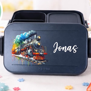 Puede incluir: Una fiambrera azul marino con una ilustración de tren colorido y el nombre "Jonas" en escritura blanca. El tren tiene un efecto de humo arcoíris. La fiambrera tiene dos compartimentos y cierres negros.