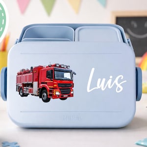 Könnte beinhalten: Hellblaue Brotdose mit rotem Feuerwehrauto-Aufkleber und dem Namen "Luis" in weißer Schrift. Die Brotdose hat einen Griff und Fächer. Ein grüner Kreis mit "Design 1" befindet sich oben links.
