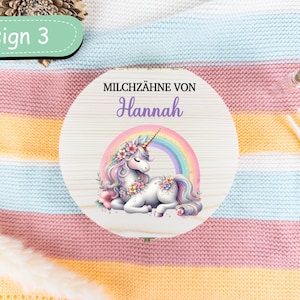 May include: A round wooden box with a unicorn illustration and the name "Hannah". The unicorn has a rainbow mane and tail, and is surrounded by a rainbow. The text "MILCHZ&Auml;HNE VON" is above the name. The box is labeled "Design 3".