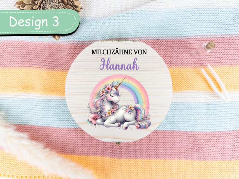 May include: A round wooden box with a unicorn illustration and the name "Hannah". The unicorn has a rainbow mane and tail, and is surrounded by a rainbow. The text "MILCHZ&Auml;HNE VON" is above the name. The box is labeled "Design 3".