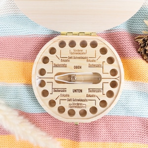 May include: A wooden baby tooth keepsake box with a circular design and multiple holes for teeth. The box has German text labeling the teeth. A clear vial is in the center. The box is on a colorful striped blanket.