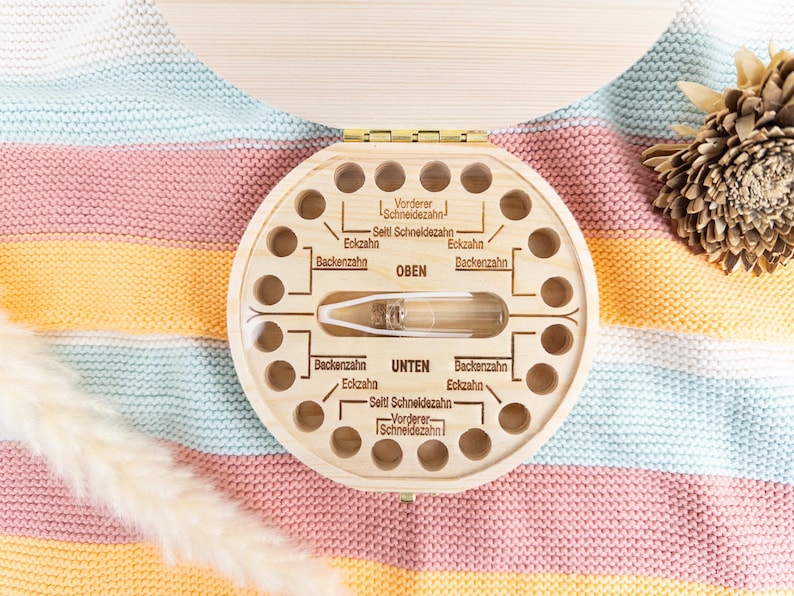 May include: A wooden baby tooth keepsake box with a circular design and multiple holes for teeth. The box has German text labeling the teeth. A clear vial is in the center. The box is on a colorful striped blanket.