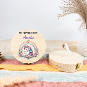 May include: A round wooden box with a hinged lid, featuring a unicorn design within a rainbow. The text "MILCHZ&Auml;HNE VON Amelie" is printed on the lid. A small glass bottle and a white vase with dried plants are also visible.