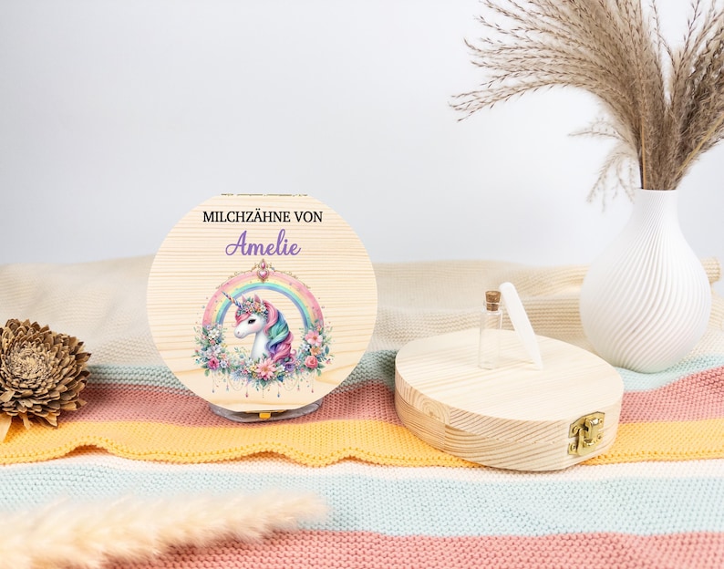 May include: A round wooden box with a hinged lid, featuring a unicorn design within a rainbow. The text "MILCHZ&Auml;HNE VON Amelie" is printed on the lid. A small glass bottle and a white vase with dried plants are also visible.