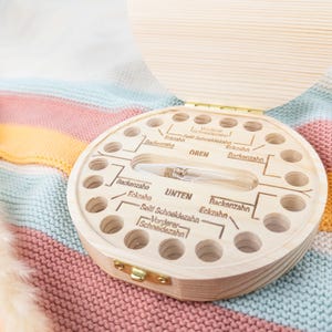 May include: A wooden baby tooth keepsake box with a hinged lid. The box is round with multiple holes and labeled sections for teeth. A small glass vial is in the center. The box is on a colorful knitted blanket.