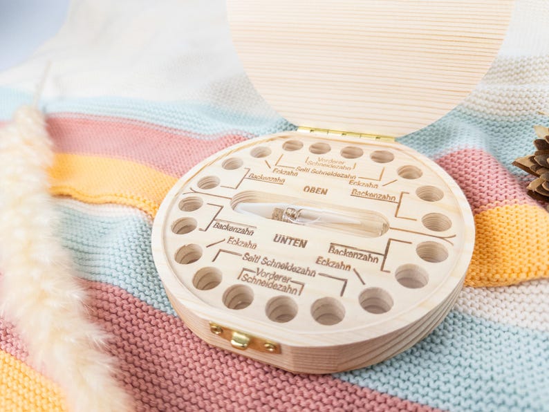 May include: A wooden baby tooth keepsake box with a hinged lid. The box is round with multiple holes and labeled sections for teeth. A small glass vial is in the center. The box is on a colorful knitted blanket.