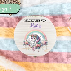 May include: A round wooden box with a unicorn design, rainbow, and the name "Malia". The box is decorated with pastel colors and floral accents. The text "Design 2" is in a green oval. The box is on a striped blanket.