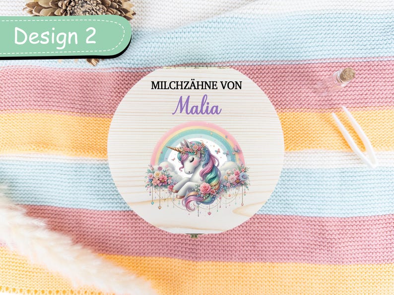 May include: A round wooden box with a unicorn design, rainbow, and the name "Malia". The box is decorated with pastel colors and floral accents. The text "Design 2" is in a green oval. The box is on a striped blanket.