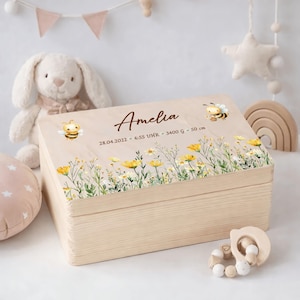 Könnte beinhalten: Eine Holzkiste mit floralem Design und dem Namen "Amelia" auf dem Deckel. Die Kiste zeigt Illustrationen von Bienen und Wildblumen. Der Text auf der Kiste lautet: 28.04.2022 - 6:55 UHR - 3400 g - 50 cm.