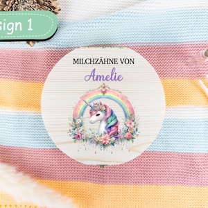 May include: A round wooden box with a unicorn design. The box features a unicorn with a rainbow, floral accents, and the name "Amelie". The text "Design 1" is in a green oval. The box is on a striped blanket.
