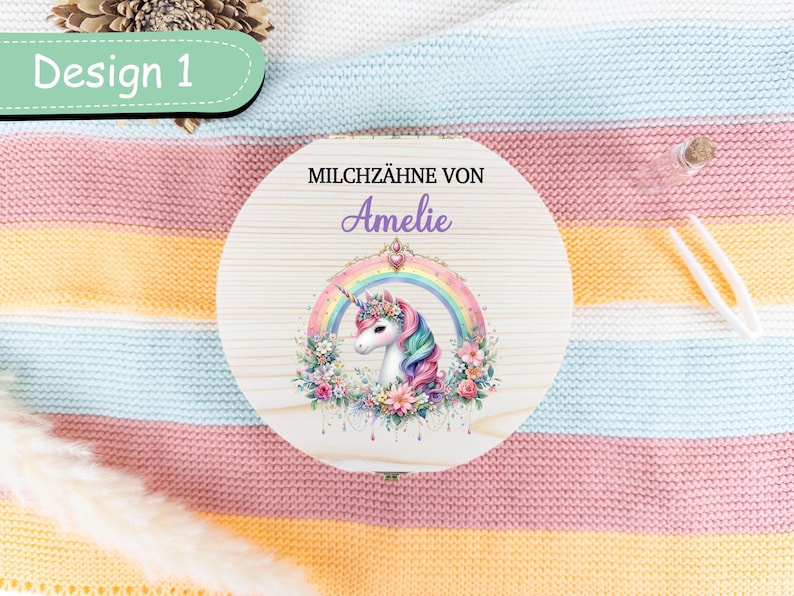 May include: A round wooden box with a unicorn design. The box features a unicorn with a rainbow, floral accents, and the name "Amelie". The text "Design 1" is in a green oval. The box is on a striped blanket.