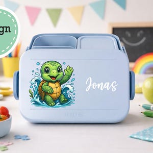 Könnte beinhalten: Hellblaue Brotdose mit einem Cartoon-Schildkröten-Design und dem Namen "Jonas" in Weiß. Die Brotdose hat einen Griff und Fächer. Das Design zeigt eine grüne Schildkröte in blauem Wasser. Schulmaterialien sind im Hintergrund.