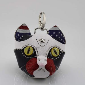 May include: A cat-shaped keychain charm featuring black, white, and red leather panels with visible stitching. The cat has yellow eyes, a small silver star on its forehead, and long clear whiskers. The inner ears display a blue and white star pattern.